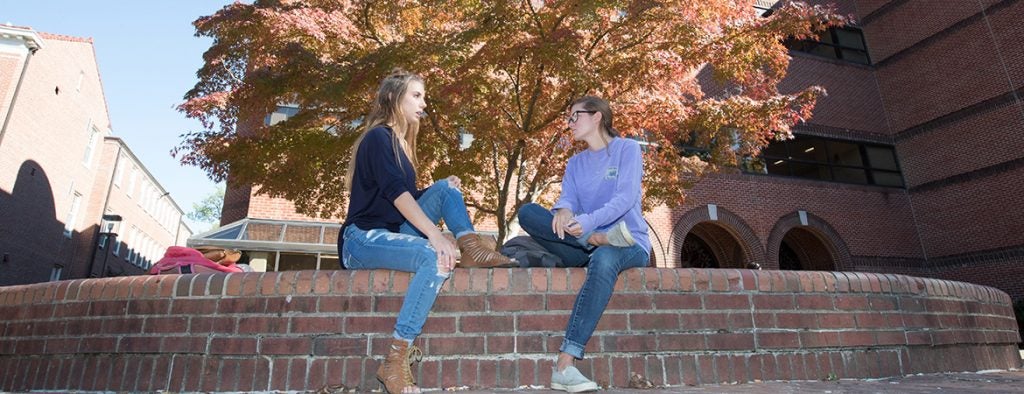 Two students sit on a brick wall facing each other.