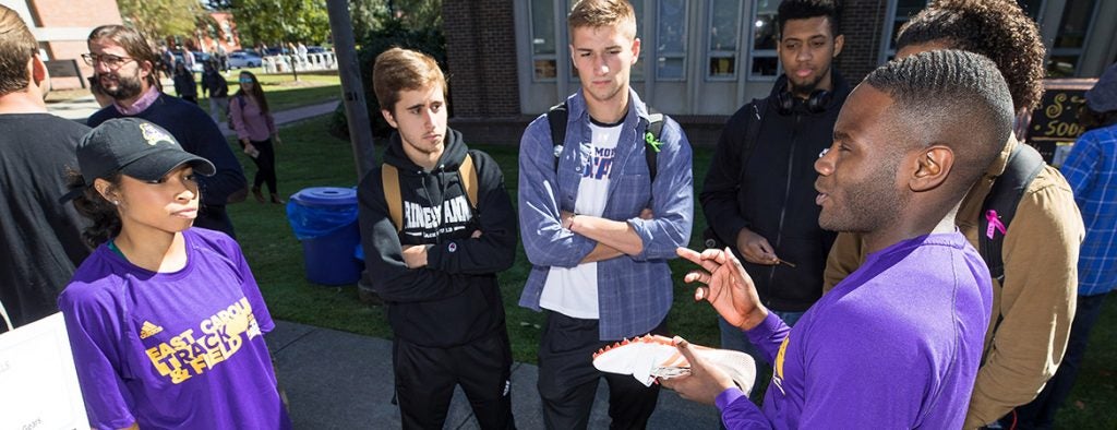 A group of students standing in a circle around a male student holding a white athletic shoe.