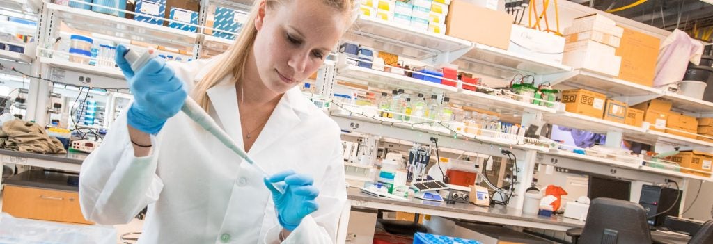 Female in white lab coat wearing gloves holds a pipet.